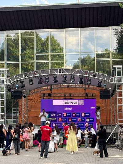 An outdoor stage set up for a Kahoot! event is the central focus of the image, located in Yerevan, Armenia. A large LED screen displays the Kahoot! logo, a game PIN of "957 7305", a QR code, and various player avatars and names. A male presenter, wearing a white shirt and red shorts, stands on the stage, addressing the audience and holding a microphone. The stage is framed by metal scaffolding and equipped with multiple spotlights and speakers. A crowd of people, some holding bags and a few with small dogs, are gathered in front of the stage, facing the screen and presenter. The background features a modern building with large glass windows reflecting trees and the sky, indicating an urban setting. The overall atmosphere suggests a public gathering or event in progress.