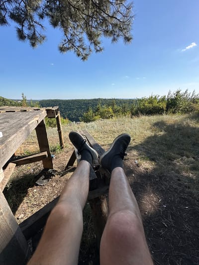A first-person perspective shot shows a person's legs and feet resting on a wooden bench. The person is wearing dark sneakers and dark socks. The background depicts a scenic overlook with a vast expanse of green forest and hills under a clear blue sky with sparse clouds. The sun is shining brightly, indicating it is daytime, likely midday or afternoon. The scene appears to be in a natural, outdoor setting, possibly a park or hiking trail. The overall mood seems relaxed and peaceful, with the person enjoying the view. There is a wooden railing in the foreground, and the ground is covered with dry grass and dirt. The location is Malozalissia, Ukraine.
