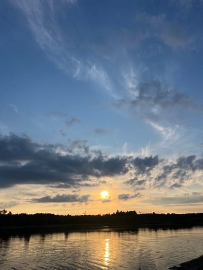 This image depicts a sunset over a body of water, likely a lake or river, in Corbetstown, Ireland. The sky is a gradient of blue transitioning to warm oranges and yellows near the horizon, where the sun is visible as a bright, round disc. Wispy white clouds are scattered across the upper sky, while darker, more substantial clouds hang lower, obscuring parts of the sunset.

Along the distant shoreline, a dense line of trees forms a dark silhouette against the illuminated sky. Several wind turbines are also visible within this treeline, their structures appearing as thin, dark lines.

The water in the foreground is calm, reflecting the colors of the sky and the silhouette of the trees. The reflection of the sun creates a shimmering, golden path across the water's surface. The overall scene is one of natural beauty and tranquility at the end of the day. No people or specific actions are depicted.