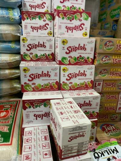 The image shows a stack of boxes of "Siplah" fruit juice in a shop in Kecamatan Lawang, Indonesia. The boxes are white with red text and feature images of various fruits. Some boxes are stacked on top of other boxes, and others are arranged in a single row. The boxes are all sealed. There are also other boxes of different brands visible behind the "Siplah" boxes. A few blue and white items are visible in the lower part of the image, but their details are unclear. The background is a wall with some printed graphics.