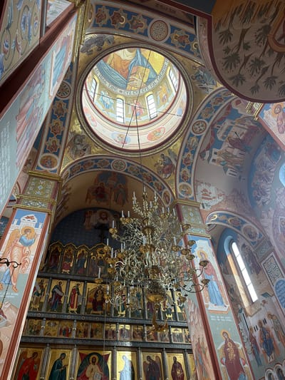 The image captures the ornate interior of the Cathedral of Christ the Saviour in Moscow, Russia, looking upwards towards the central dome and ceiling. The architecture features arched ceilings adorned with vibrant murals depicting biblical scenes and figures in traditional Orthodox Christian iconography. A large, elaborate golden chandelier hangs from the ceiling, casting a warm light on the scene. Sunlight streams in from high windows, illuminating the richly decorated walls and dome. The walls are covered in religious paintings and mosaics, with intricate patterns and gold leaf details. A section of an iconostasis, a wall of religious icons, is visible in the lower part of the image, showcasing rows of framed religious artwork. The overall impression is one of grandeur, solemnity, and religious devotion.