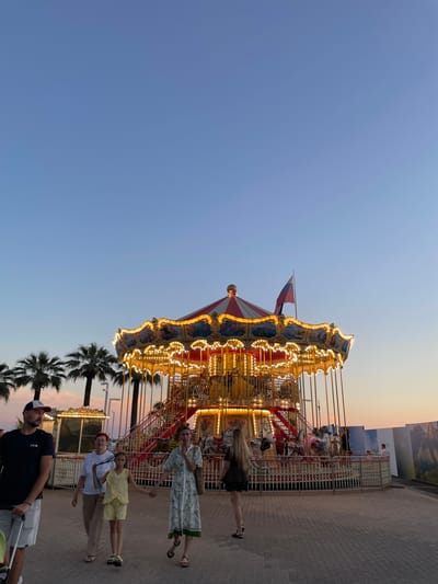 In Sochi, Russia, a brightly lit carousel stands as the central focus of this image. The carousel, adorned with numerous lights, appears to be in operation, with people visible on and around it. The scene is set against a backdrop of a clear, pale blue sky transitioning to a warm, orange-tinged sunset. Several palm trees are visible, suggesting a warm climate and potentially a seaside location.
A group of people are captured in the frame. In the foreground, a man wearing a baseball cap and t-shirt is pushing a stroller. To his right, a woman in a white top and beige pants, and a young girl in a yellow dress are walking. Further to the right, another woman in a patterned dress is walking, holding hands with a younger girl. The people appear to be enjoying a leisurely evening outing.
The carousel itself is elaborately decorated with paintings and ornate trim, illuminated by a multitude of warm yellow lights that create a festive atmosphere. A French flag is visible flying from the carousel structure. The ground around the carousel is paved with brickwork. The overall mood is relaxed and pleasant, indicative of an evening at an amusement or entertainment venue during pleasant weather.