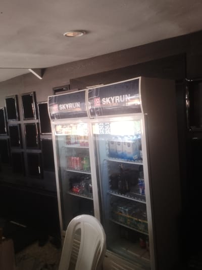 The image shows two refrigerated display cases filled with beverages, likely in a store or bar setting in Howayi, Nigeria. The display cases are branded with the "SKYRUN" logo. One case is packed with bottles of what appear to be soft drinks and juices, while the other contains bottles of water and some canned beverages, possibly energy drinks. The shelves are organized, and the products are clearly visible through the glass doors.
In the foreground, a white plastic chair is partially visible, suggesting a place where customers might sit. The background features a wall with several framed square panels, possibly mirrors or decorative elements, adding to the interior design of the establishment. The ceiling has a recessed light fixture. The lighting suggests it could be daytime, with artificial light from inside the refrigerators and potentially overhead lighting in the establishment. The overall scene depicts a retail environment stocked and ready for business. There is no visible indication of people actively engaged in any activity, though the presence of a chair implies a customer-facing area.