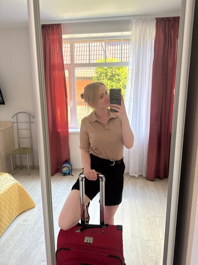 The image shows a mirror reflection of a woman holding a smartphone and taking a selfie. She is wearing a beige collared shirt and black shorts with a black belt. She is kneeling next to a red suitcase. The scene appears to be indoors, possibly a hotel room, with a window in the background showing trees and tiled rooftops, suggesting a daytime setting. The room has wooden floors and is decorated with red and white curtains. A small backpack with cartoon characters is visible on the floor. The reflection also captures a metal garment rack and a yellow quilted cushion. The woman's expression is neutral. There is no visible text in the image.