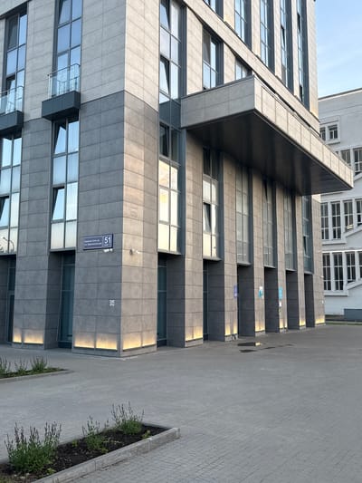 A low-angle, daytime shot of the exterior of a gray, modern office building. The building is located at 51 Ul. Krasnosel'skaya in Kazan, Russia. The building features a prominent overhang and numerous windows with some windows slightly ajar. The building's facade is constructed of gray panels. The sidewalk is made of gray paving stones and features narrow garden beds with small, flowering plants. Another modern office building is located in the background, painted white. There are a few visible security cameras on the building.