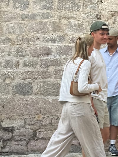 In Budva, Montenegro, three individuals are captured in a candid moment against a textured stone wall. A woman in the foreground, with blonde hair pulled back, wears a white cable-knit sweater and loose, light-colored linen pants. She carries a small, tan shoulder bag. To her right, a young man sporting a green baseball cap and a white t-shirt, smiles as he looks off to the side. Partially visible behind him is another man wearing a light blue collared shirt and a light-colored cap, also looking away. The scene is likely a street in Budva's Old Town, characterized by its ancient stone architecture.