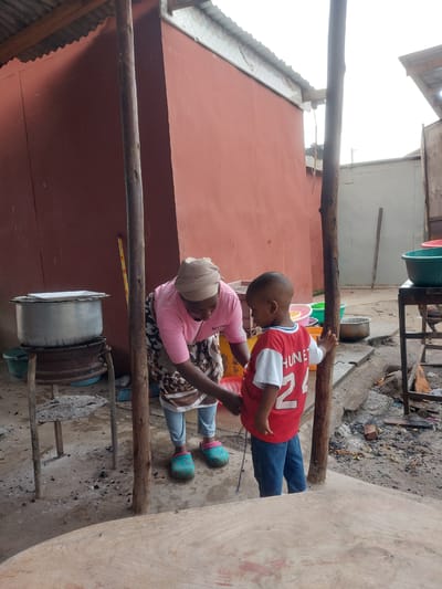 The image shows a child in a red shirt with the number 22, and blue jeans, next to a woman wearing a pink shirt, a patterned skirt, and a head covering. They are both standing next to a wooden pole that seems to form part of a makeshift structure. Beside the woman is a large metal pot on a rudimentary stove. The ground appears to be a dirt surface with scattered debris and ash. In the background, there is a red building and other structures indicating a densely populated area, potentially suggesting an urban setting in Dar es-Salaam, Tanzania. The interaction between the child and woman suggests a familial connection.