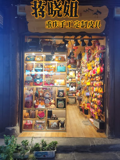 The image shows the front of a shop in the Shapingba District, China. A vertical wooden sign with illuminated Chinese characters stands to the left of the shop window, advertising handmade leather goods. The shop window displays a variety of handmade leather products arranged in a wooden shelving unit. On the top of the unit, colorful cartoon character toys are displayed. Below them, leather bags and purses fill the shelves, some decorated with traditional motifs. Below the shop window, two planters are placed with greenery in them.