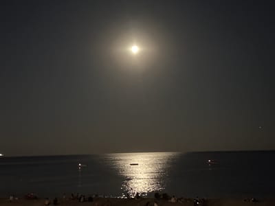 The image depicts a nocturnal scene at a beach in Antalya, Turkey. The moon is prominently visible in the sky, casting a bright, shimmering reflection on the surface of the sea. The sea itself appears calm, with the exception of the moonlight's effect. In the distance, there are a few boats on the water, some with visible lights. On the beach in the foreground, a number of people are gathered, some sitting and others standing. They are silhouetted against the dim light. The overall atmosphere of the image is peaceful and serene, suggesting a quiet evening by the sea.