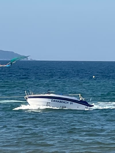 A white speed boat with "DANABEACH 01" printed on its side is moving through blue water, leaving a white wake. The boat has a dark blue stripe along its upper edge and a metal rail on top. In the background, there is a vast expanse of blue ocean under a clear, light blue sky. To the left, partially visible, is another boat with a green sail or canopy. In the far distance, a hazy, mountainous coastline is visible. A small yellow buoy floats on the water to the right of the speed boat. The scene is set on a bright, sunny day, likely in the waters off Đà Nẵng, Vietnam, given the boat's labeling.