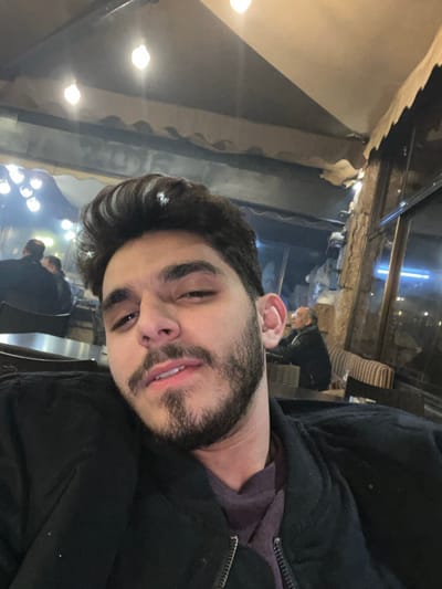 A man with dark hair and a beard is posing for a selfie in what appears to be a restaurant. He's wearing a dark jacket and a purple shirt. In the background, there are other patrons and soft lighting. The restaurant seems to have a unique interior design with visible ceiling structures and a slightly worn appearance. The lighting suggests it could be nighttime.