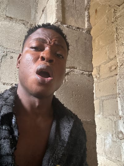 A young Black male is captured in a close-up, low-angle selfie. He has dark, short hair and his face is angled towards the camera, with his mouth open. He is wearing a black garment with a textured, woven appearance and fringed edges. The background consists of textured concrete walls, suggesting an urban or industrial setting. The lighting appears to be natural and somewhat harsh, casting shadows on his face and the walls. The image does not contain any visual cues to specifically place it within Ikorodu, Nigeria.