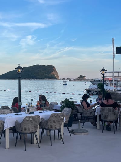 The image depicts an outdoor seating area of a restaurant or cafe in Budva, Montenegro, overlooking the Adriatic Sea. In the foreground, several tables are set with white tablecloths and chairs. People are seated at some of these tables, engaged in dining and conversation. One table has a woman in a black dress and a young boy in a plaid shirt. Another table has three women, one in a red top and two in dark attire. A street lamp stands prominently in the left mid-ground.
In the background, the calm sea is visible, with a large, green, mountainous island dominating the horizon. Several boats are moored or moving in the water. The sky is a clear blue with scattered, wispy clouds, suggesting it is late afternoon or early evening. The overall scene conveys a relaxed, social atmosphere.