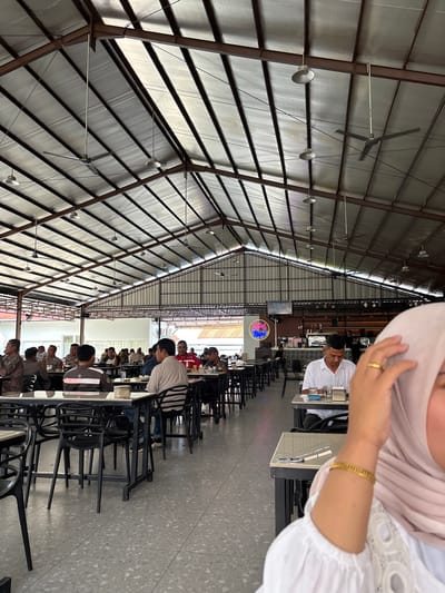 The image depicts the interior of a spacious, open-air restaurant in Bireuen, Indonesia. The ceiling is high and constructed with metal sheeting and beams, featuring multiple industrial-style ceiling fans and hanging lights. The floor is a terrazzo material. Several tables with black chairs are arranged throughout the space, occupied by patrons. In the foreground, a woman wearing a light pink hijab and a white top is partially visible, with her right hand touching her head and adorned with a gold ring and bracelet. In the mid-ground, a man in a white shirt is seated at a table, looking down. Further back, various groups of men are seated at tables, some engaged in conversation. In the background, a counter area with a coffee machine and a neon sign that reads "SPA" is visible. The overall atmosphere suggests a casual dining environment.