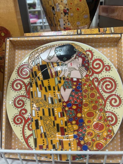 This media file shows a decorative plate featuring a depiction of "The Kiss" by Gustav Klimt. The plate is adorned with the iconic imagery of a couple embracing, rendered in vibrant gold, yellow, red, and blue tones, characteristic of Klimt's Art Nouveau style. The couple is intertwined in a golden robe, with the man kissing the woman on the cheek. The background of the plate is a pale yellow with scattered white dots, and a red and gold swirling border frames the central image.
The plate is presented within a light brown box that has a pattern of yellow polka dots on a light brown background. The box also features some of the same decorative elements seen on the plate, with gold and red swirling patterns. Partially visible to the left of the plate is another object, possibly a matching item or part of the packaging, also adorned with Klimt's distinctive golden circular motifs.
The overall impression is of a decorative collectible item, likely intended for display. The lighting suggests an indoor setting, possibly a retail environment, given the presence of a white wire shelving unit on the right side of the image. No specific time of day, weather, or emotional cues are discernible beyond the romantic subject matter of the artwork. There is no visible text in the media file.
