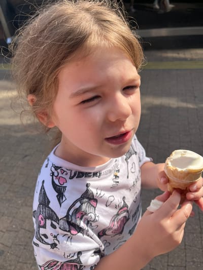 The media file shows a close-up, rotated image of a young girl outdoors, holding an ice cream cone. The girl, with blonde hair, is wearing a white t-shirt with a cartoon print of cat-like characters in black and pink. Her eyes are closed, and her mouth is slightly open as she holds the ice cream. Her hands are visible, one holding the cone and the other holding a small white piece of tissue. The background appears to be a paved surface, possibly a sidewalk or pathway. The lighting suggests it is daytime, with sunlight casting shadows. The girl's expression appears content or perhaps experiencing the cold of the ice cream. The overall scene evokes a casual, summery moment. The location context is Sochi, Russia. There is no visible text in the image.