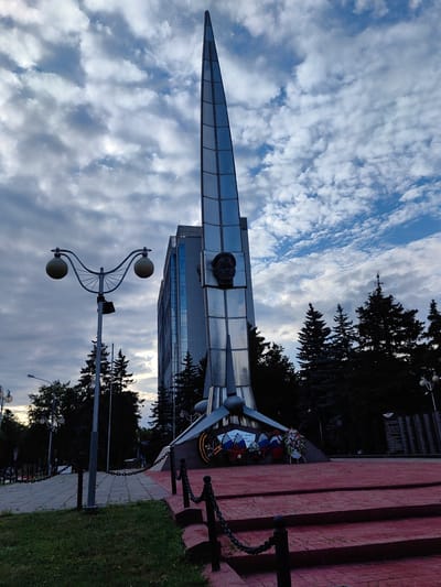 This media file captures a monument in Vnukovo, Russia, against a dramatic sky filled with white and grey clouds. The monument, tall and sharp, resembles a rocket or a spire and is made of reflective material. At its base, several wreaths are laid, suggesting a memorial or commemoration. A bronze bust of a man is prominently featured on the monument.
In the foreground, a red paved area with steps leads up to the monument, bordered by dark metal chains and posts. To the left, a distinctive street lamp with two round lights stands on a pole. Behind the monument, a modern glass-fronted building rises, contrasting with the monument's design. Evergreen trees form a dark silhouette against the sky. The overall mood is solemn and contemplative, with the cloudy sky adding a sense of gravitas. The time of day appears to be late afternoon or early evening, given the lighting and cloud cover.
On a plaque at the base of the monument, some Russian text is visible, including "ПЕР. ГЕРОЯМ АВИАЦИИ" and "1941-1945 гг.", indicating a tribute to aviation heroes from the period of 1941-1945. To the right, another dark board displays text and an emblem, likely listing names or commemorating an event.