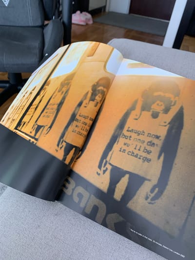 An open book or magazine is depicted at an angle, displaying two pages featuring stencil artwork. The primary artwork, visible on the right page, shows a black silhouette of a figure, possibly a child, holding a sign against an orange-brown background. The sign contains the text: "Laugh now, but one day we'll be in charge." A similar, partially visible stencil of another figure with the same text on a sign is seen on the left page.

Along the bottom right edge of the page, white text provides context for the artwork: "You're no safer in first class. 20 minutes, District Line, London 2002." Part of the artist's name, "Bane" (likely referring to Banksy), is also visible on the right page.

The book's pages have a glossy finish, reflecting light. The setting appears to be indoors, with a light gray upholstered surface visible to the right of the book, possibly part of a chair or sofa. Darker furniture or objects and a glimpse of a wooden floor are visible in the background on the left. The image is a static shot of the open publication.