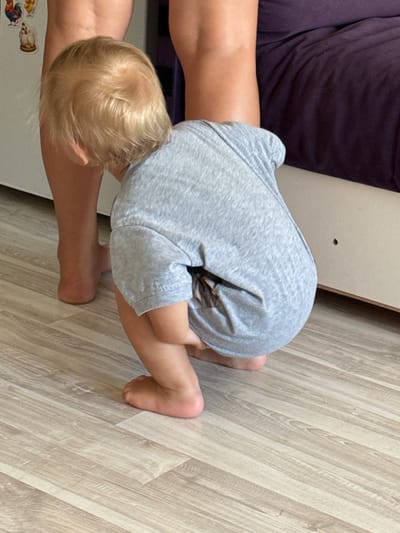 A blonde-haired toddler, facing away from the camera and towards a person's legs, is squatting on a light-colored laminate floor. The toddler is wearing a gray t-shirt. The legs in front of the toddler are bare and appear to be from an adult. In the background, a dark purple upholstered surface is visible, possibly a couch or bed, and a white cabinet with decorative chicken magnets. The scene is indoors, with the lighting suggesting daylight. The location is not identifiable as Sochi, Russia, or any specific city.