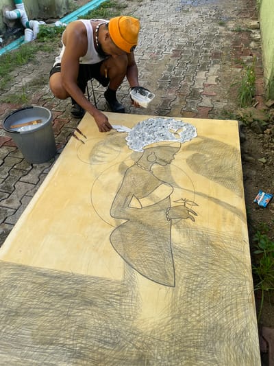 A man wearing an orange beanie, a white tank top, and black shorts is squatting next to a large rectangular board with a drawing of a woman. The woman in the drawing is in profile, facing to the left, wearing a large afro and jewelry. The drawing is partially painted white, particularly the afro. The man is holding a small container in his left hand and a tool in his right hand, which he is using to apply the white paint to the drawing.

The scene is outdoors, on a brick-paved surface. A gray bucket with a paintbrush inside is next to the drawing board. A discarded wrapper is on the ground near the board. White PVC pipes are visible on the left side of the frame. The location is Ado, Nigeria.