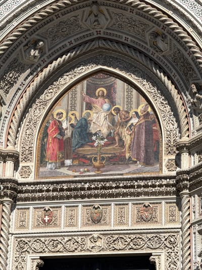 This image captures a detailed section of the central portal on the main facade of the Cattedrale di Santa Maria del Fiore (Florence Cathedral) in Florence, Italy. The prominent feature is a large, arched mosaic depicting a religious scene.
In the mosaic, Jesus Christ is centrally depicted, enthroned, with a golden halo, raising his right hand and holding a blue orb in his left hand, wearing a pinkish-red robe. To the viewer's left of Christ, a figure in a red robe holds a palm frond, standing beside a kneeling figure. On the floor below them, a golden vase contains white lilies. To the viewer's right of Christ, several figures are present, including one bare-chested individual being led or presented, and another robed figure with a halo. At the bottom edge of the mosaic, text visible on a dark band reads "VENEZIA 1886 SOCIETÀ MVSIVA VEN", indicating its origin and date.
The mosaic is framed by an elaborate architectural archway crafted from light-colored marble with darker decorative inlays. This archway features multiple concentric bands of intricate carvings, including twisted columnar elements, floral motifs, geometric patterns, and relief sculptures of figures, some holding books. Below the mosaic and above the unseen entrance, there are rectangular panels containing heraldic shields, one prominently displaying a red cross on a white field. These panels are set against a background of small mosaic tesserae and surrounded by further detailed stone carvings. Flanking the entire composition are spiraling columns with ornate capitals. The scene is illuminated by natural daylight.
