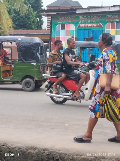 The image captures a street scene in Aba, Nigeria, likely on a commercial street given the presence of shops and vehicles. In the foreground, a woman walks away from the camera, wearing a colorful, floral-patterned dress and black sandals, carrying a beige purse and a yellow and black striped bag. She is walking on a sidewalk made of a rough, textured material. To her left, a red motorcycle with a white panel is ridden by two men. The man in front wears a black t-shirt and grey shorts, while the man behind him wears a vibrantly patterned short-sleeved shirt and dark shorts. The motorcycle is positioned on the asphalt road. Behind the motorcycle, a green auto-rickshaw, also known as a "keke," is parked or moving slowly. The auto-rickshaw has a black tarp covering its passenger area, and part of some writing in English is visible on its side, which appears to read "SUS WEPT." Further in the background, a building with a colorful facade displays signage for "GMC PAINT DEP" and a phone number, "08139099717". The overall environment suggests an active urban setting with typical street traffic and commerce. Palm trees and other foliage are visible in the upper left corner, indicating a tropical climate. The date and time stamp "06/26/2026 17:11" is present at the bottom right, and "REDMI 15" is at the bottom left.