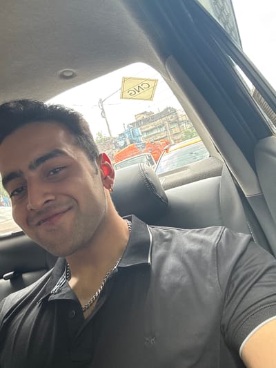 A man is seated in a car in Kolkata, India, facing the camera. He has dark hair, dark eyes, and fair skin, and is wearing a dark gray polo shirt with a silver chain around his neck. He appears to be smiling subtly. The car interior is gray, with the roof and seat visible. Outside the car window, buildings and street scenes of Kolkata are visible, including a CNG sign. The day is bright, and the image is taken from a slightly angled perspective.