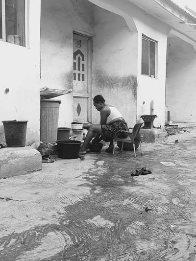 The black and white image captures a scene in Lokoja, Nigeria. A woman is seated on a chair, leaning forward with her hands inside a bucket. The setting appears to be an outdoor courtyard or alleyway adjacent to a building. There are several buckets and containers arranged in a line, suggesting the area is used for washing or cleaning. The building's wall features a door with geometric designs, a window, and what appears to be textured concrete. The ground is uneven, potentially concrete, and shows signs of wear. A pair of shoes is discarded next to the chair.