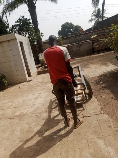 The scene is set outdoors in Akwanga, Nigeria, under bright sunlight, indicated by long shadows cast on the ground. A male individual, viewed from behind, is centered in the frame, actively pushing a two-wheeled, dark-colored metal handcart. He is dressed in a red t-shirt with white sleeves, dark brown trousers, and light brown slip-on sandals. The handcart contains at least one light-colored, possibly white, bag.
The ground immediately around the individual is paved with light-colored, square concrete tiles, which transitions to a rougher, lighter-toned dirt or gravel surface towards the right side of the image.
To the left of the individual, a distinct, light-colored, rectangular structure, likely tiled in white, stands with a visible wooden door. A small potted plant is situated near its base. Further left and towards the top, a tall palm tree with a rough trunk is visible, alongside other green foliage.
In the mid-ground, a low concrete wall stretches horizontally, topped with various materials including corrugated metal sheets and wooden planks. Adjacent to this wall, a white Intermediate Bulk Container (IBC) tank, appearing partially filled with a green liquid, is present. Behind the wall, additional trees and parts of distant structures are discernible. Overhead, multiple power lines traverse the clear, bright sky.