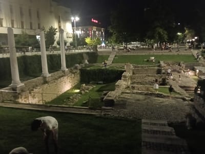 The image depicts a night scene at the Roman Stadium of Philippopolis in Plovdiv, Bulgaria. The stadium, an archaeological site, is visible with its ancient stone ruins and tiered seating areas. In the foreground, a person in a white shirt is bent over on a grassy lawn. In the midground, several people are scattered around the ruins and walkways, some appearing to be tourists. White stone pillars stand prominently on the left side of the image. In the background, a modern building with illuminated windows and signage is visible. The overall atmosphere is urban and public, with the site being a popular attraction after dark.