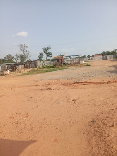 The image shows an outdoor scene in Tasha, Nigeria. The foreground is a dirt road with tire tracks and scattered debris. In the middle ground, there are several structures that appear to be temporary shelters or construction sheds, made from corrugated metal and wood. Some of these structures have tarpaulin roofs. There is also a pile of gravel and a concrete wall under construction. Trees with sparse foliage are visible in the background, along with a power line. The sky is clear with a few scattered clouds. The overall impression is of an undeveloped or under-construction area.