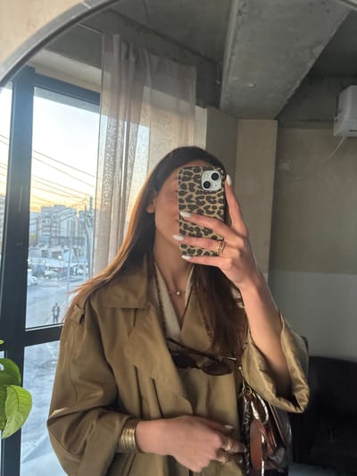 A person is taking a mirror selfie with a smartphone with a leopard-print case. The person is wearing a beige trench coat and jewelry including rings and bracelets. Sunglasses are partially visible near the person's chest. Through the window, an urban cityscape is visible with buildings, roads, and vehicles. A plant with green leaves is at the bottom of the frame. The reflection suggests the photo is taken in an indoor space. There is no evidence to indicate the location is in Zovuni, Armenia.
