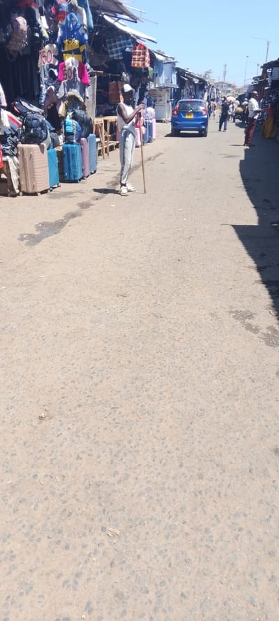 The image captures a vibrant street market scene in Nairobi, Kenya, under clear, sunny skies. A wide, dusty asphalt road forms the central path, flanked on both sides by numerous market stalls. These stalls appear to be makeshift structures with corrugated iron roofs and wooden supports, displaying a wide variety of goods.

In the foreground, slightly to the left, a man stands facing towards the right. He wears a white sleeveless top, light-colored track pants with dark stripes, white sneakers, and a white cap, holding a long wooden stick. Behind him, along the left side of the road, numerous items of clothing, including jackets and shirts, are hung on display. Below these, a row of travel luggage, including large suitcases in shades of tan, blue, and pink, are stacked on the ground. Price tags with "150" and "50" are visible on some hanging garments, likely indicating prices in Kenyan Shillings.

Further down the road, a blue hatchback car is visible, moving or parked. Beyond the car, the street continues into the distance, lined with more stalls and bustling with people walking and conducting business. Shadows are cast sharply on the right side of the road and near the stalls, indicating a bright daytime setting, likely mid-morning or early afternoon. The overall scene depicts a typical busy day in a lively urban market environment.