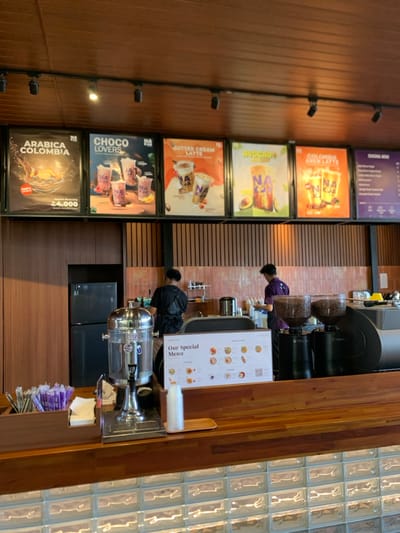 The scene is a cafe located at the coordinates -7.957, 112.622, in the city of Malang, Indonesia. Two men are standing behind the counter. One is facing the camera, wearing a black shirt, the other is facing away from the camera, wearing a purple shirt. There are several glass containers behind the counter with unknown liquids in them, along with a black, stainless steel coffee grinder. On the counter, there is a large, stainless steel container with clear liquid and a "Our Special Menu" sign. There are two smaller stainless steel coffee grinders and a large, stainless steel espresso machine. There is a large, black, metal refrigerator behind the counter. Five framed posters are on the wall behind the counter, each advertising different types of coffee or tea. The counter is made of wood. Below the counter there is a glass brick wall. There are some drinking straws in a container next to the counter.