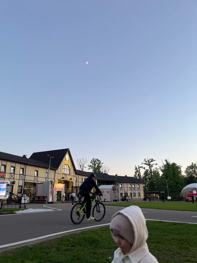 The image depicts a street scene in Odintsovo, Russia, at dusk. A cyclist is performing a trick on a path, while a young child wearing a hooded jacket observes the scene from the side. The building behind them has a clock and is decorated with lights, which are also on a structure on the side. The path is bordered by a grass area on the right. In the background are trees and a building with a dome. The sky is clear, with a crescent moon visible.