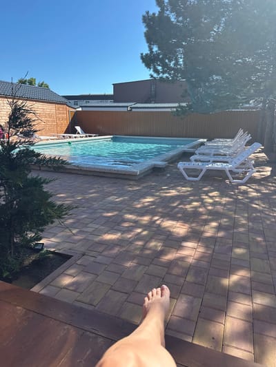 The image depicts a sunny day at an outdoor swimming pool area. The pool is filled with clear blue water and surrounded by a paved patio. Several white lounge chairs are arranged neatly on the patio, facing the pool. A wooden fence encloses the area, and some trees and buildings are visible in the background. A person's bare foot is prominently featured in the foreground, suggesting they are relaxing by the pool. The scene appears to be a resort or a private residence in Anapa, Russia, given the context.