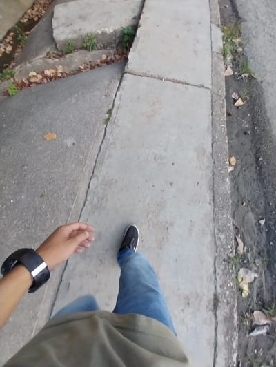 A first-person, high-angle view depicts a person walking on a concrete sidewalk. The person is wearing blue jeans and black shoes. A black smartwatch is visible on the left wrist. The sidewalk is bordered by a curb with grass and leaves. The location is Tinaquillo, Venezuela.