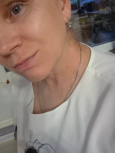 The media file is a close-up, slightly tilted, portrait of a woman. She is wearing a white top with a delicate silver chain necklace. Her face is partially visible, with her lips and a portion of her eye in focus. She has light skin and blue eyes. A small, delicate earring with a light blue gem is visible on her right ear. The background is blurred, but it appears to be an indoor setting with some natural light, possibly near a window. The overall impression is casual and personal. There is no discernible text in the image. The lighting suggests it could be daytime.