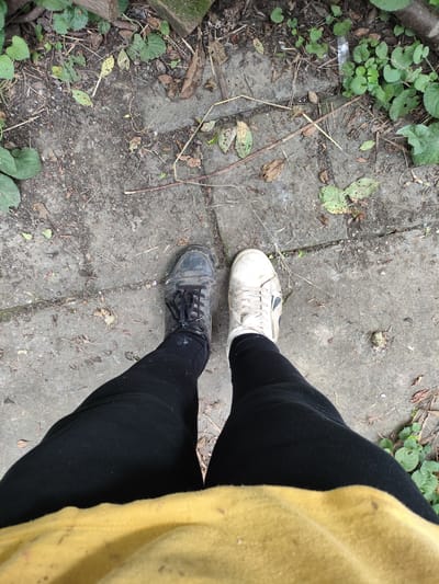 An overhead, full-body shot depicts a person standing on a concrete path with patches of dirt and scattered leaves. The person is wearing black leggings and a yellow top, with a pair of dark, laced sneakers on their left foot and white, laced sneakers on their right foot. The context of Znamenskiy, Russia is not evident from the image.