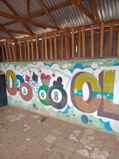 The image shows a mural painted on a wall in Durumi, Nigeria. The mural depicts playing cards, dice, and pool balls, with the word "COOL" written in large, stylized letters. To the left of the mural, there is a black door or entrance. The wall itself is painted with a light green background and blue and white cloud-like shapes. Above the mural, the wooden structure of a roof with corrugated metal roofing is visible, supported by wooden beams and rafters. The floor in front of the mural is covered with irregularly shaped, light brown tiles. A small bird with dark and grey feathers is perched on the tiled floor in the lower right portion of the image. The overall scene appears to be an outdoor covered area.