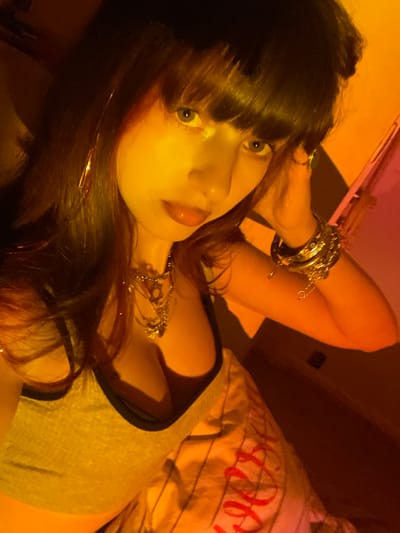 A woman with dark hair and bangs gazes at the camera. She is lying down, wearing a gray tank top with black trim and a layered silver necklace. Her arms are raised, adorned with numerous silver bracelets. The warm lighting casts a golden hue over the scene, with a striped pillow visible in the background. The composition is tilted, adding to the casual, intimate feel of the image.