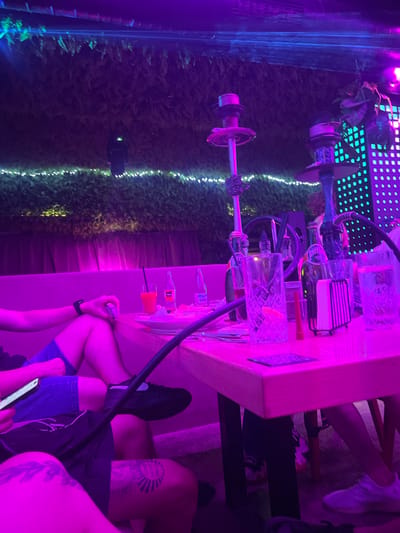 The image depicts a dimly lit, vibrant interior, heavily illuminated by pink and purple neon lighting. The scene appears to be a lounge or bar area, possibly a shisha bar, given the presence of two hookahs prominently featured in the mid-ground. The hookahs are tall, elaborate apparatuses with stems, bowls, and hoses.

A wooden table sits in the foreground, covered with various items. These include glasses, bottles, a napkin holder with napkins, and what seems to be food on a plate. One glass contains a red beverage with a straw, and another contains ice. A phone is visible in someone's hand resting on the table.

Several people are present, though their faces are not clearly visible due to the lighting and framing of the shot. We see legs and arms, suggesting they are seated around the table. One person is wearing blue shorts and a black sneaker, while another has on dark shorts and a white sneaker. Tattoos are visible on the legs of at least one individual. The flooring is not clearly discernible.

The background features a wall covered with lush green foliage, possibly artificial plants or ivy, illuminated by soft white string lights. Further back, a wall of blue LED lights arranged in a grid pattern is visible, adding to the overall ambiance of the establishment. The overall impression is one of a modern, stylish, and possibly trendy nightspot. Given the context, this establishment is located in Sofia, Bulgaria.