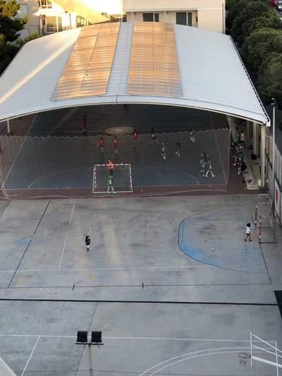 The image shows an outdoor sports court with a covered section featuring solar panels on the roof, located in the city of València, Spain. The court appears to be a futsal or basketball court, with painted lines and markings on the concrete surface. There are two small goals visible on the court. In the covered area, a group of young people, dressed in athletic wear, are playing futsal. One individual in a green and white uniform is in goal. Several other individuals, some in red and white uniforms, are scattered on the court. Outside the covered area, a single individual in a white shirt and black shorts is walking across the court. There are also spectators sitting on a bench in the covered area. In the background, a building with multiple stories is visible, along with trees and a street. The lighting suggests it is either early morning or late afternoon, with sunlight casting long shadows.