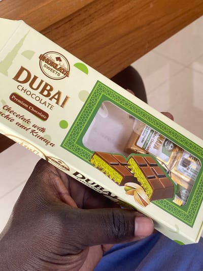 A hand is holding a box of "Dubai Chocolate" in Kediri, Indonesia. The box is cream-colored with green and gold accents. It features an image of the Burj Khalifa and stylized patterns. The text on the box includes "DUBAI CHOCOLATE," "Premium Chocolate," and "Chocolate with Pistachio and Kunafa." Inside the box, through a clear window, several individually wrapped chocolate pieces are visible. The background appears to be a wooden surface, possibly a table, and a blue fabric.