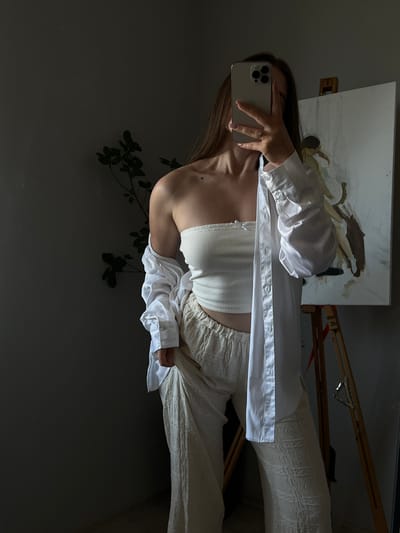The image captures a person taking a mirror selfie in what appears to be a room with an easel and a painting. The person is wearing a white ribbed tube top and a white unbuttoned oversized shirt, with cream-colored pants. Their right hand is holding a smartphone, and a ring is visible on their left hand. Behind them, a painting is propped on an easel. The scene is dimly lit, suggesting it might be evening or the room has limited natural light. A plant with dark green leaves is visible in the foreground, casting shadows on the dark wall. The overall mood seems relaxed and personal, typical of a casual selfie. The image is oriented vertically, with the top of the person's head towards the left side of the frame. There is no visible text in the image.