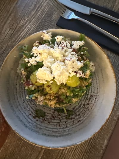 A detailed overhead view presents a freshly prepared salad served in a ceramic bowl on a dark wooden table, located in Plovdiv, Bulgaria. The salad consists of a base of finely chopped green vegetables and grains, topped with a generous mound of crumbled white cheese. Several green grapes are nestled within the cheese and greens. A reddish-brown spice, consistent with sumac, is visibly sprinkled over the cheese and across the textured, light grey interior of the bowl. The bowl features a slightly irregular, earthy-toned rim and subtle concentric ridges. To the upper right of the bowl, a polished silver fork and knife are neatly placed on a dark, rectangular placemat. The wooden table exhibits a pronounced grain and texture, suggesting a rustic setting. No individuals are present in the frame.