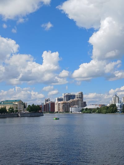 This media file depicts a scenic view of a city skyline alongside a body of water, likely a river, on a bright, cloudy day. The scene is oriented vertically, suggesting the original photo was taken in portrait mode or has been rotated.
On the left side of the image, a vibrant blue sky is filled with large, puffy white clouds. The right side of the frame showcases a cityscape with a variety of buildings, ranging from modern high-rises to older structures, lining the waterfront. Lush green trees are interspersed among the buildings, providing a natural contrast to the urban architecture.
The water is calm, with gentle ripples on its surface, reflecting the sky and surrounding structures. A small, bright green boat is visible on the water, adding a touch of color and suggesting movement or activity. Along the edge of the water, there's a concrete embankment or promenade, with some pedestrians possibly present, though they are not clearly discernible.
The overall atmosphere appears peaceful and pleasant, indicative of a daytime scene. The lighting suggests it is likely late morning or afternoon. The presence of diverse architectural styles hints at a city with a history and ongoing development. While specific location cues within the image are limited to the general urban and waterfront setting, the provided context of Yekaterinburg, Russia, helps place the scene geographically. No discernible text is present in the image.
