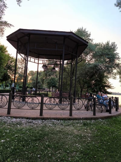 The image depicts a park scene with a large, dark-colored hexagonal gazebo at its center. The gazebo has a metal roof supported by dark pillars. Inside and around the gazebo, several people are seated on benches. In the foreground, a decorative metal fence with circular motifs separates the paved area from a grassy lawn littered with white fluffy material, possibly from cottonwood trees. Beyond the gazebo, trees and foliage are visible, with a hint of a body of water in the background, suggesting a park situated near a river or lake. The lighting suggests it is either early morning or late evening, with the sun casting a warm glow through the trees. A few bicycles are parked near the fence. The overall impression is a tranquil park setting.
