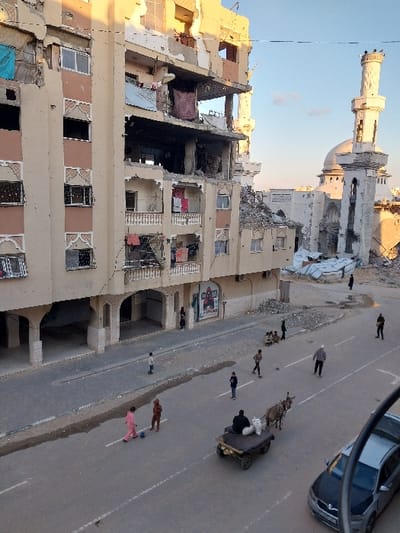 The image depicts a street scene in Khan Yunis, Palestinian Territory, during late afternoon or early morning. A multi-story residential building on the left shows significant structural damage, with upper floors partially collapsed, exposed interiors, missing windows, and damaged balconies. Debris is visible within and around the structure. To the right, a mosque with a distinct minaret and dome is also damaged, with rubble present at its base and a partially destroyed minaret top. The paved street below features white lane markings and patches of debris or moisture. Numerous individuals populate the street: two children in pink and brown clothing play with a ball in the road, a man sits on a donkey-drawn cart moving away from the viewer, and several other adults and children walk or stand along the street. A silver sedan is partially visible on the far right. The scene is illuminated by warm, low-angle sunlight, casting long shadows.