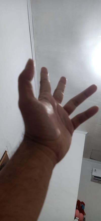 An over-the-shoulder, first-person perspective shot shows a person's left hand, with fingers spread, reaching upwards against a white wall and ceiling in what appears to be an indoor environment. The hand is in focus, while the background, including a white wall, ceiling with trim, an air conditioning unit, and some stuffed toys, is out of focus. The lighting suggests a bright, possibly artificial light source coming from the upper right. This image was taken on August 15, 2023, in Lhokseumawe, Indonesia.